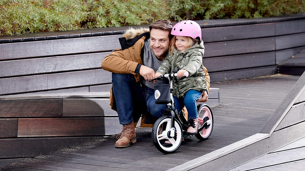 Il papà segue sua figlia che va su una bicicletta senza pedali. Ha in testa un casco rosa, scende dalla collinetta e ride tanto