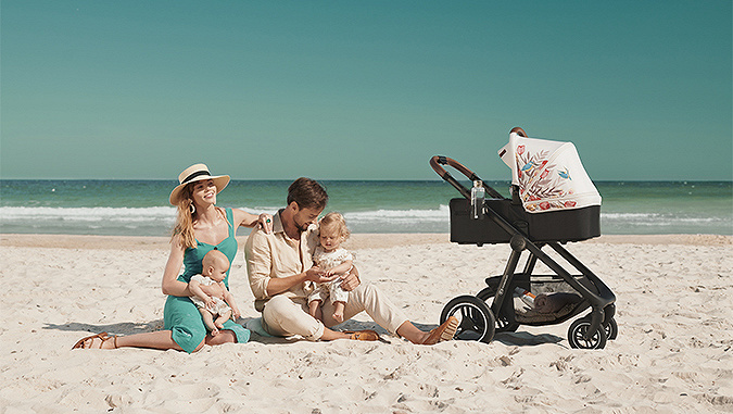In una giornata di sole, i genitori si siedono in riva al mare con i loro due bambini piccoli. Accanto a loro c'è la carrozzina - la navicella di Kinderkraft con la cappottina colorata.