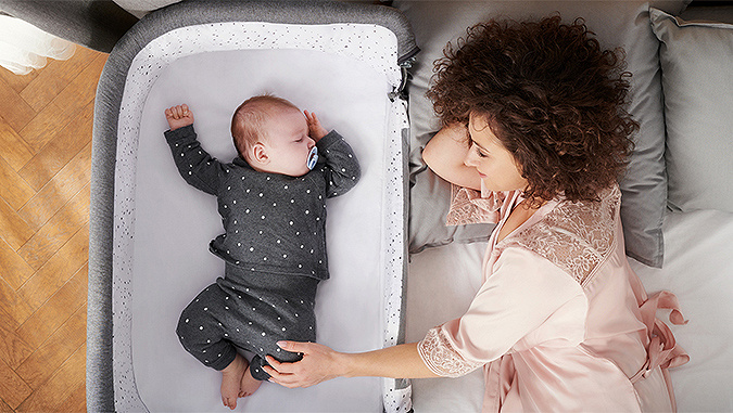 In cameretta, sdraiata sul letto, la mamma alza lo sguardo con un sorriso e tiene per un piedino il piccolo bambino, che le dorme accanto nella culla di Kinderkraft.