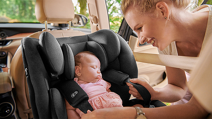 Il neonato dorme in macchina su un seggiolino auto grigio di Kinderkraft, rivolto nel senso di marcia, e la madre sorridente lo allaccia con le cinture di sicurezza.