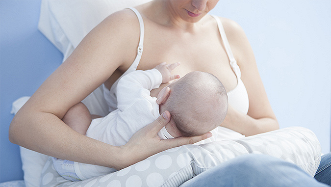 La mamma è seduta sul letto con la schiena contro il muro e allatta il neonato sdraiato sullo speciale cuscino da allattamento di Kinderkraft.
