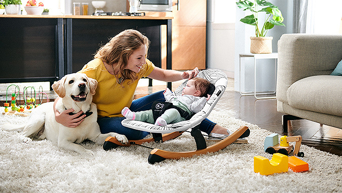 A casa, la mamma è seduta sul tappeto e accarezza il suo bambino piccolo sdraiato sulla sdraietta a dondolo di Kinderkraft. L'altra mano della mamma abbraccia il cane.