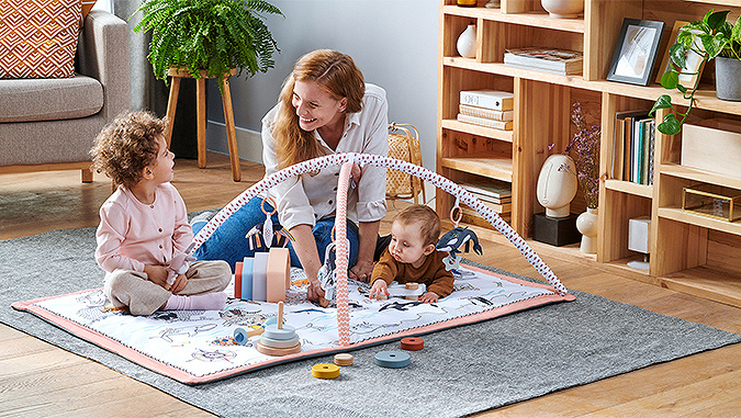 Nell'appartamento, sul pavimento è steso il tappetino da gioco di Kinderkraft. Si divertono la mamma, il ragazzino e il neonato.