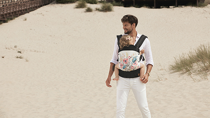 Un papà elegantemente vestito con una camicia bianca e pantaloni, sta in piedi sulla spiaggia. Sulla sua pancia nel marsupio di Kinderkraft si trova la bambina.