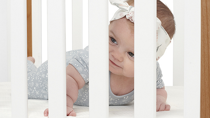 Il bambino di pochi mesi sdraiato sul comodo materassino guarda attraverso i pioli bianchi del lettino di Kinderkraft.