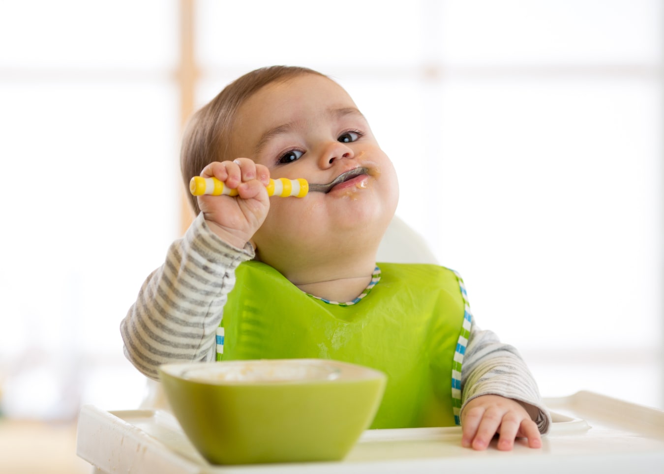 Il bambino è seduto sul seggiolone da pappa con un piattino verde davanti a lui, indossa un bavaglino verde. Mangia con il cucchiaio e guarda la telecamera.