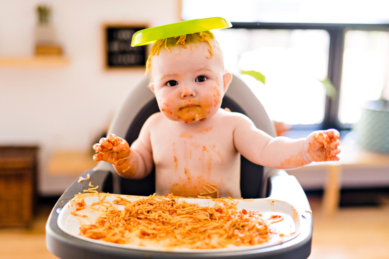 Un bambino, sporco di spaghetti, è seduto su un seggiolone con un piatto con del cibo sulla testa.