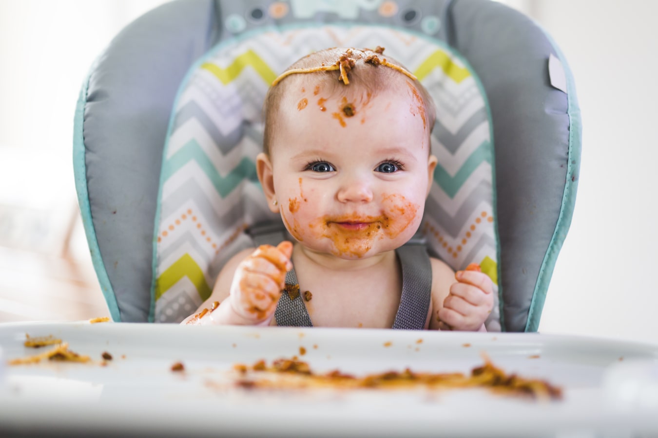 Il bambino, sporco di spaghetti, è seduto su un seggiolone. È felice e sorridente, e c'è molto cibo sia su di lui che sul vassoio.