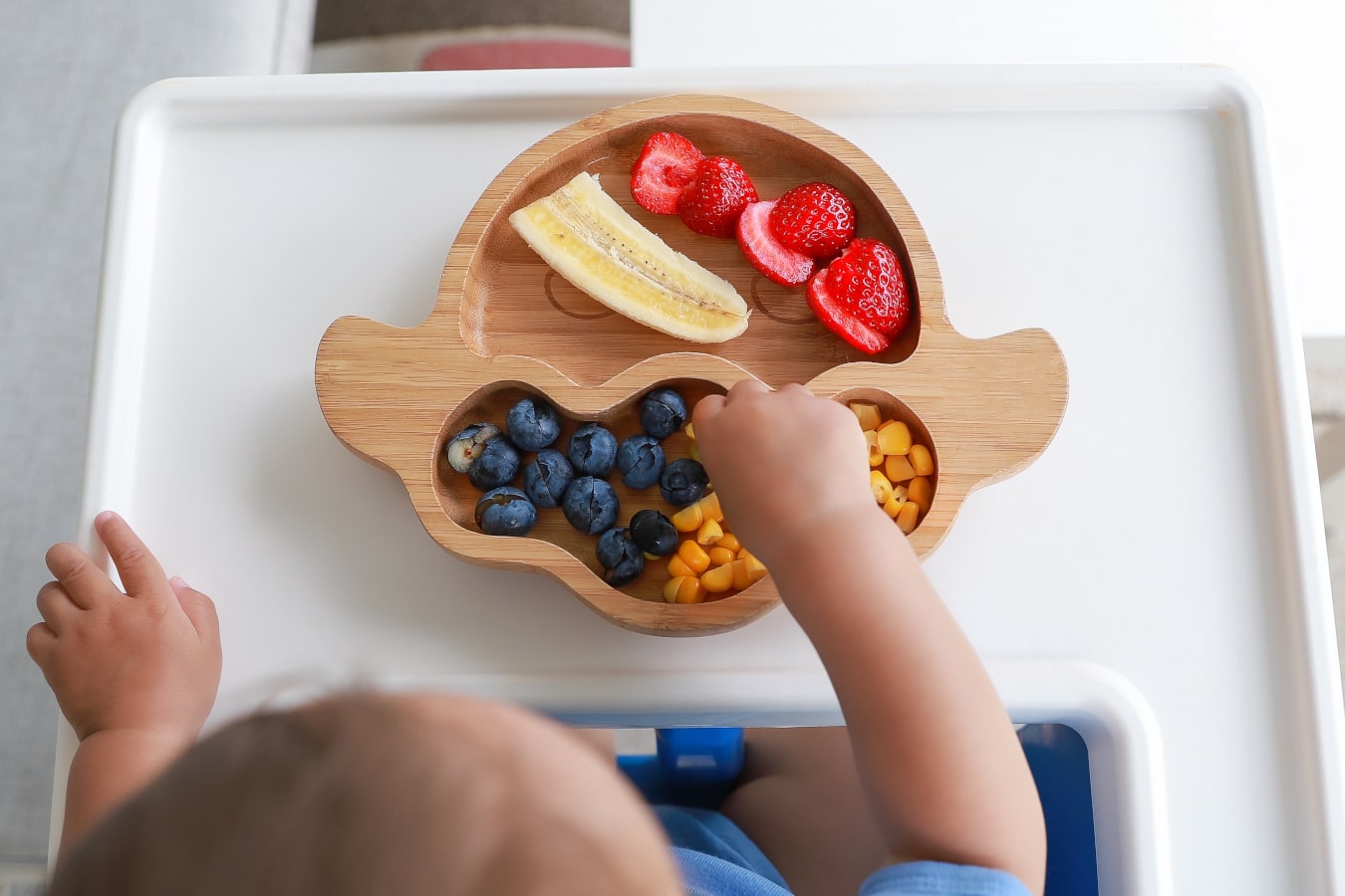 Su un piatto di legno con divisori, si può vedere la frutta e verdura multicolori: banane, mirtilli, mais e fragole. Il bambino prende il cibo.
