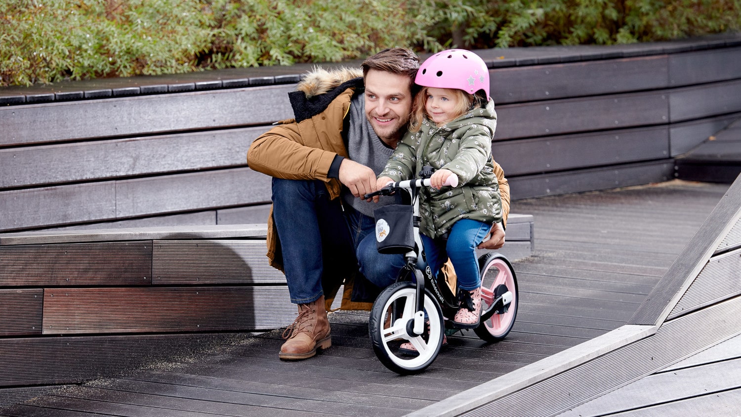 In autunno, all`aperto, il papà si accovaccia accanto alla ragazza seduta sulla bicicletta senza pedali in colore nero e rosa che indossa il casco. 