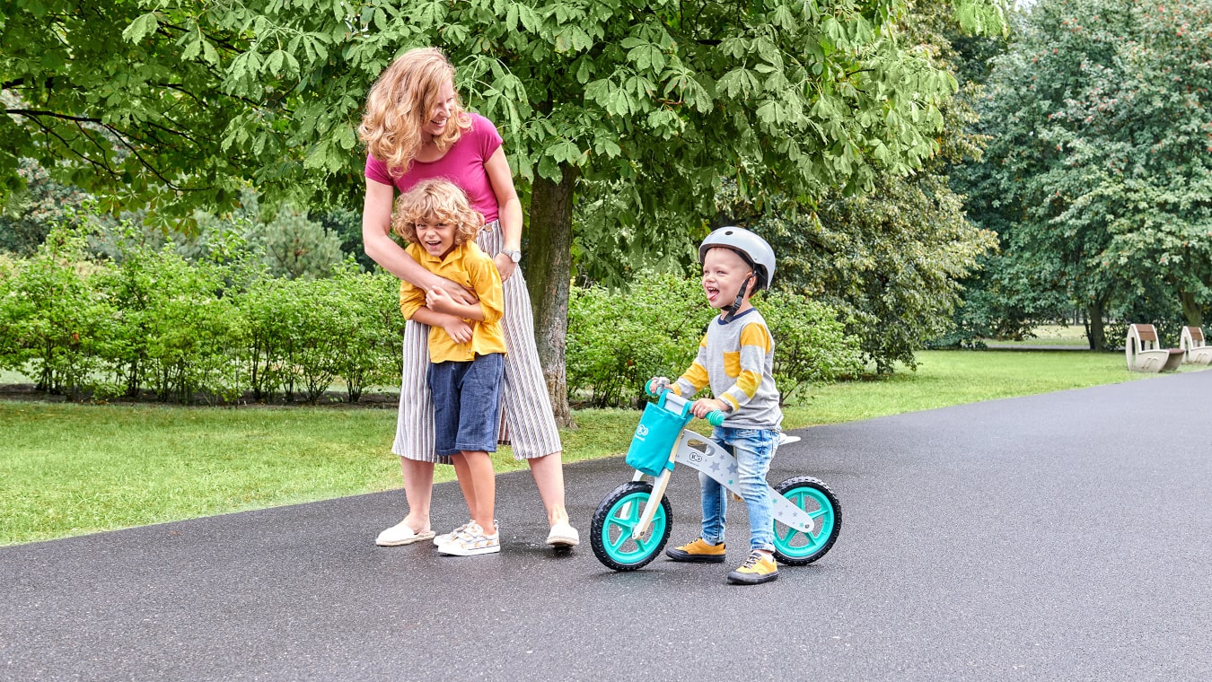 La mamma sta passeggiando nei giardini pubblici con un bambino sulla bicicletta senza pedali di color turchese e con l'altro figlio più grande