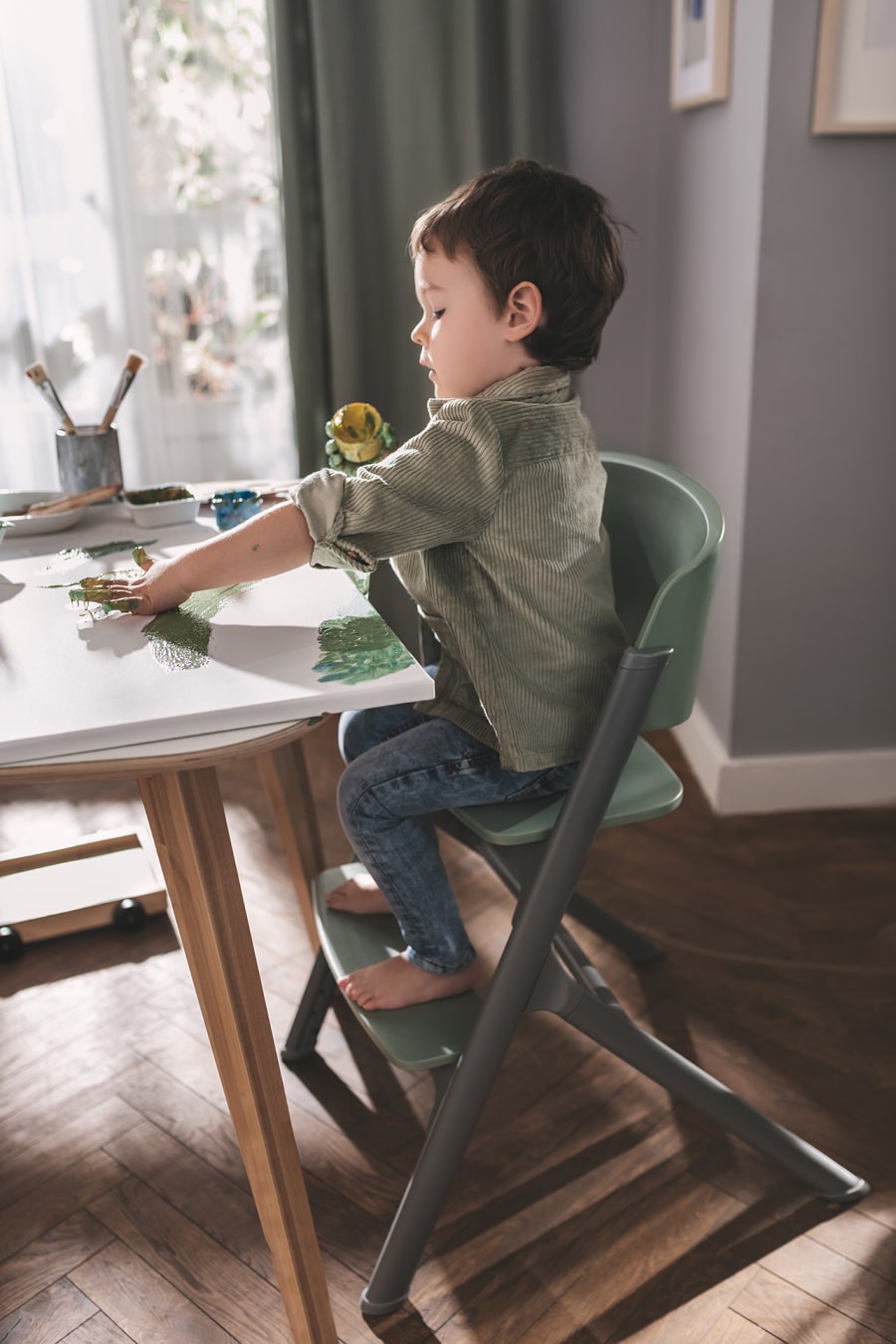 Un bambino è seduto sul seggiolone da pappa LIVY di  Kinderkraft, dipinge sulla tela con le dita, è concentrato e rilassato