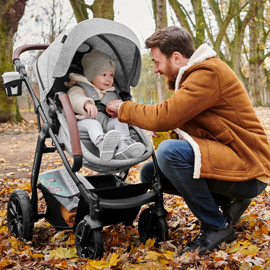 Il papà che indossa la giacca si china sul bambino seduto sul passeggino. Il bambino sta ridendo, intorno ci sono le foglie autunnali.