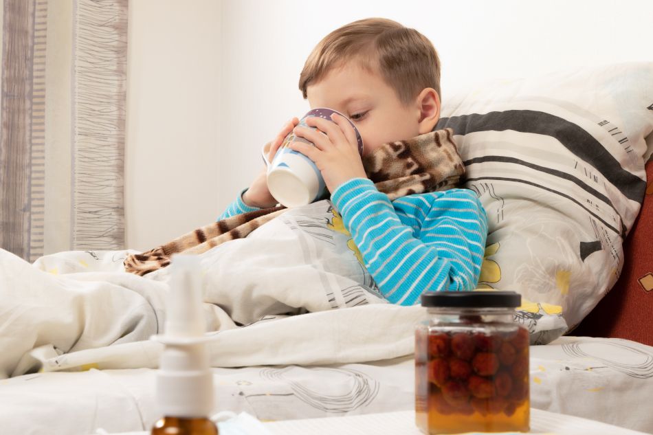Un ragazzo malato beve il tè caldo dalla tazza. Sul tavolo c'è la bottiglia con lo sciroppo di lamponi, ideale per il raffreddore.