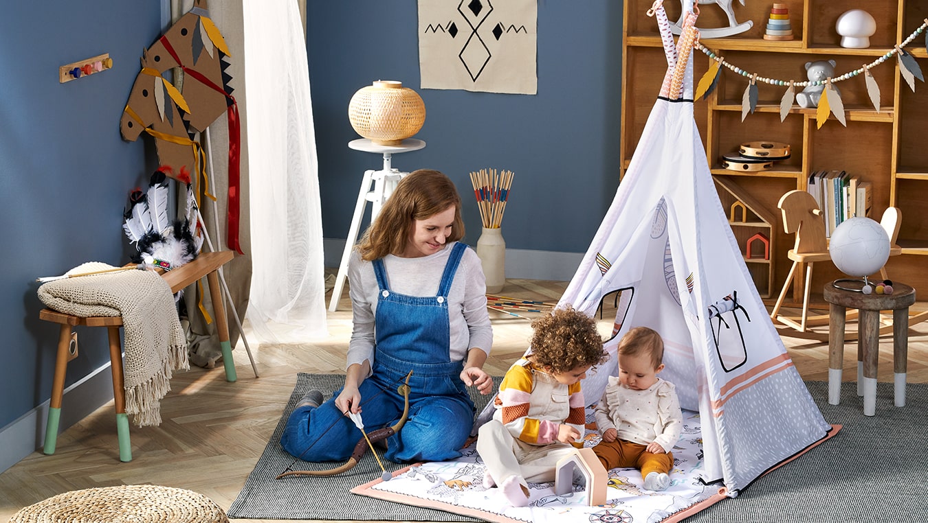Due bambini giocano nella tenda tippy di Kinderkraft, la mamma è seduta accanto e tiene in mano un arco giocattolo, tutti si divertono e ridono. 