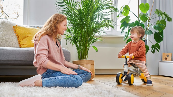 Mamma sorride verso il figlio, che gira in triciclo cutie di Kinderkraft. Sono a casa, dietro si vede delle piante verdi e un divano grigio. Il bambino sorride.
