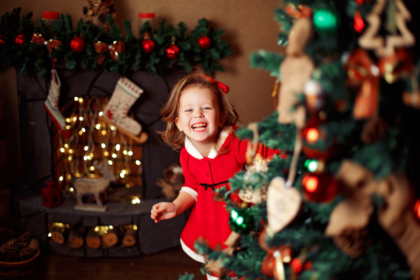 La ragazza allegra con un vestito rosso guarda nascosta dietro l'albero di Natale, ride ed è molto felice.